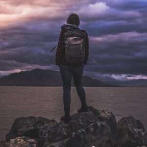 Wanderer mit Rucksack auf einem Felsen am Wasser unter Abendhimmel